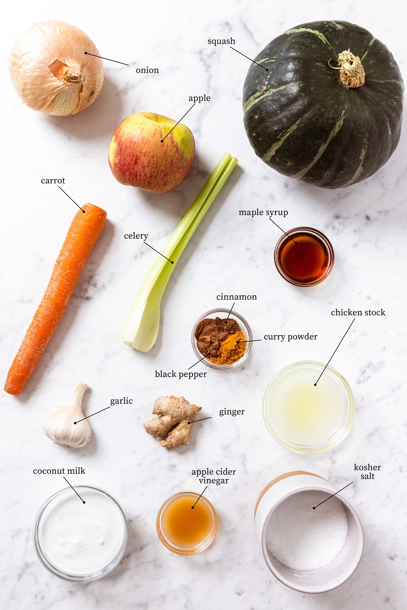 White marble surface filled with all ingredients needed to make a slow cooker squash soup including squash, apple, celery, carrot, spices, chicken stock, and more.