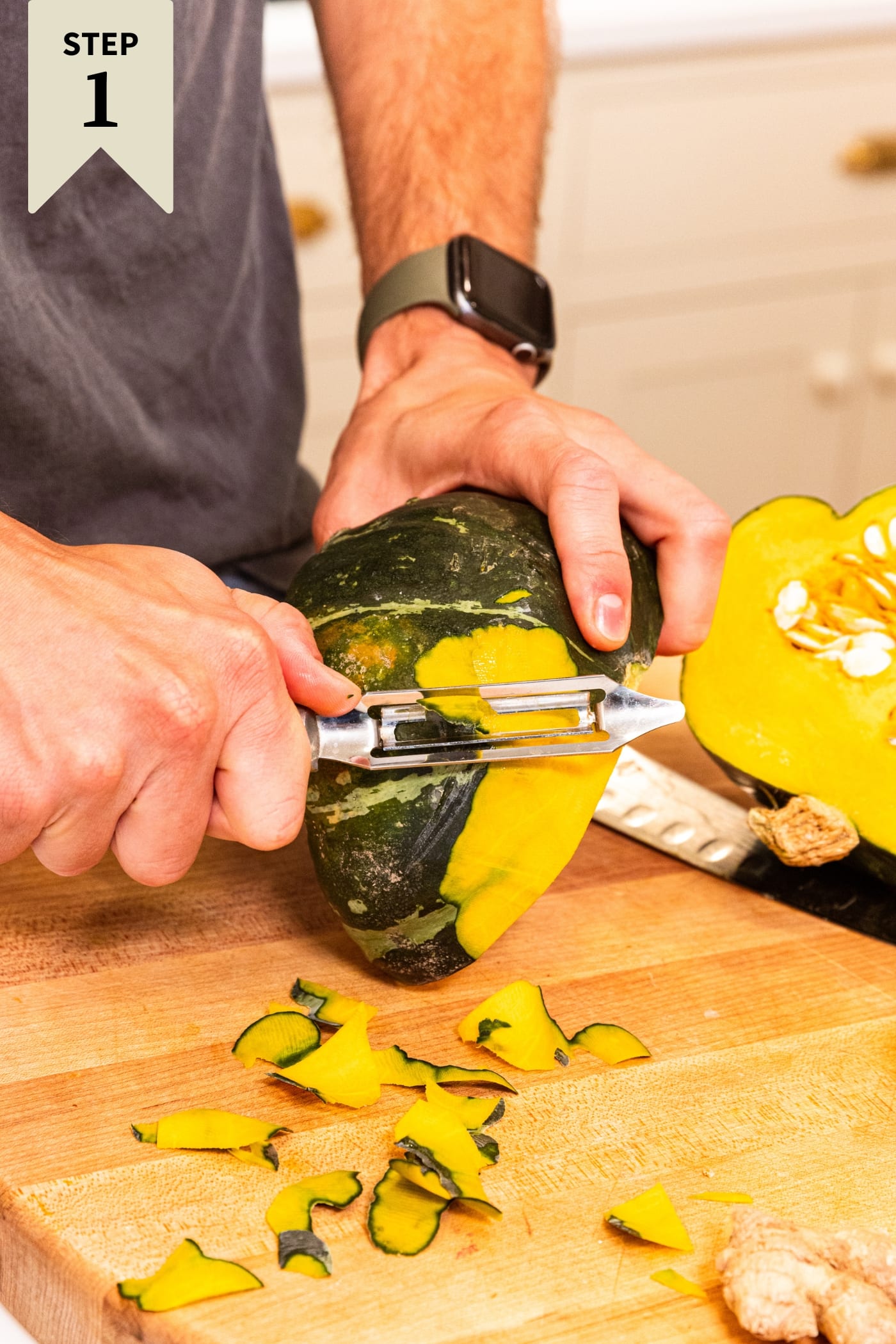 Hand holding squash using a vegetable peeler to peel a round squash from its green flesh on a wood cutting board.