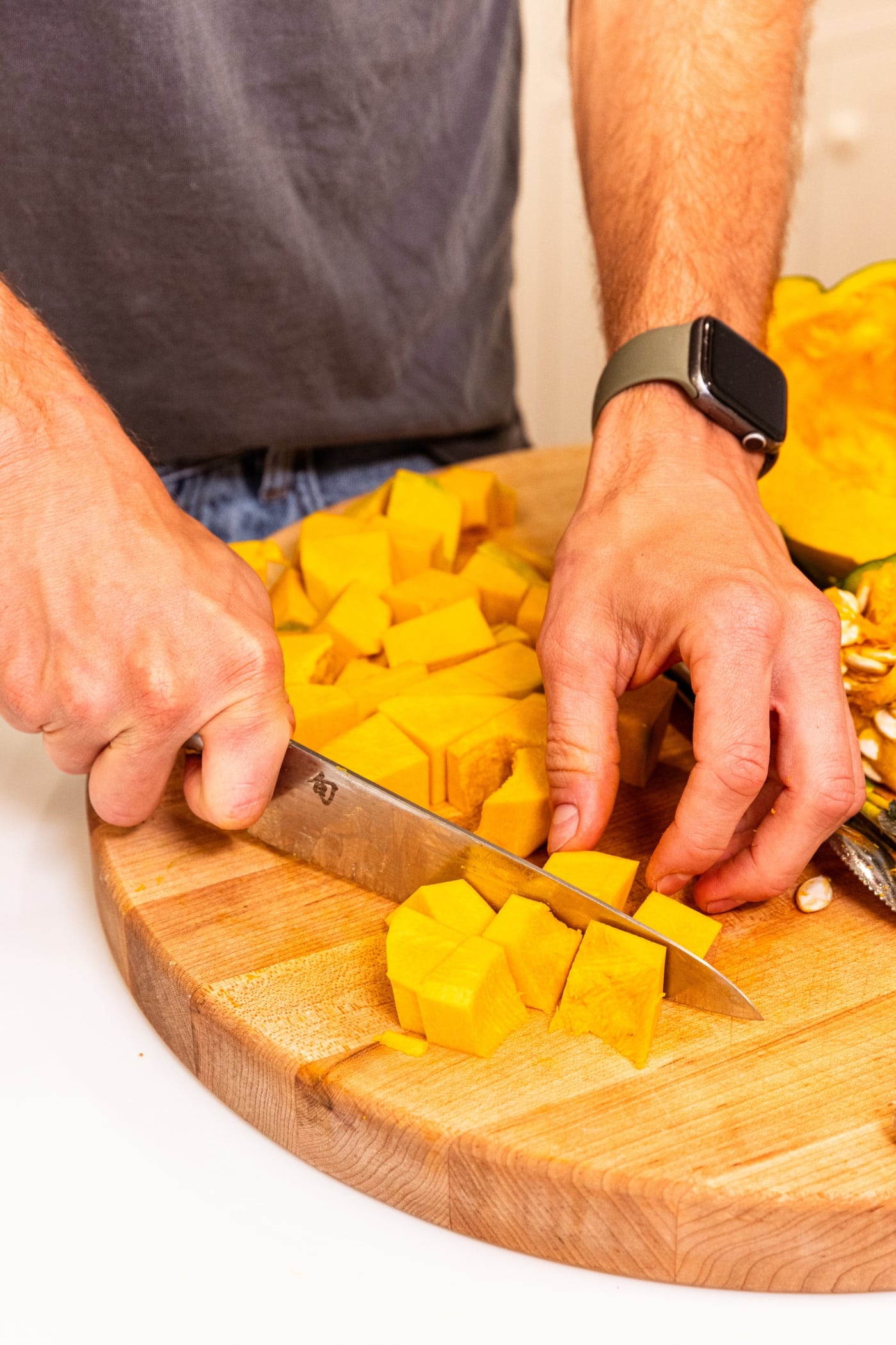 Hands using knife to slice orange colored squash into squash pieces on wood cutting board.