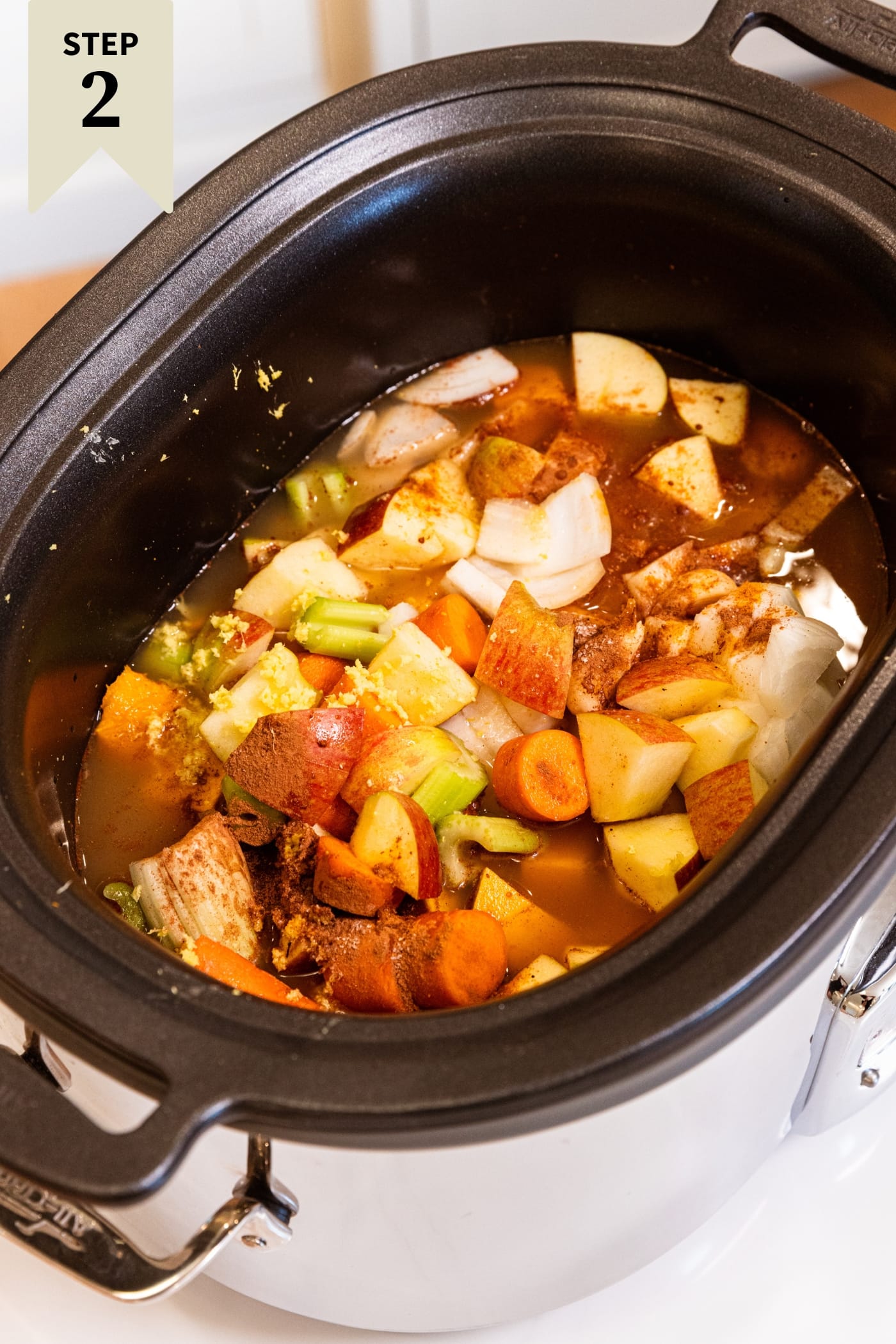 Slow cooker filled with pieces of squash, apples, carrots, chicken stock, and more before being cooked.
