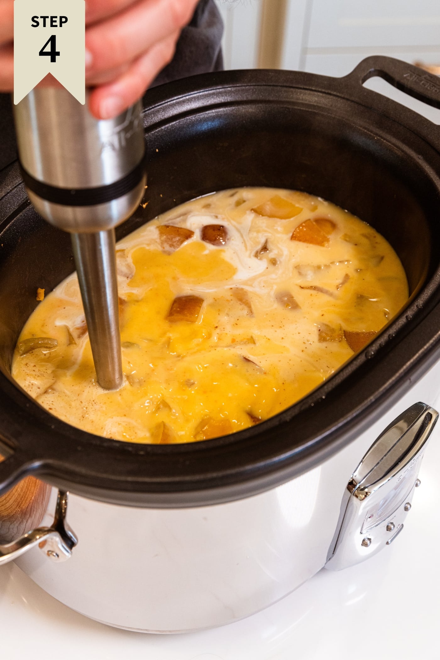 Hand holding immersion blender in a large slow cooker soup blending everything together including apples and squash.
