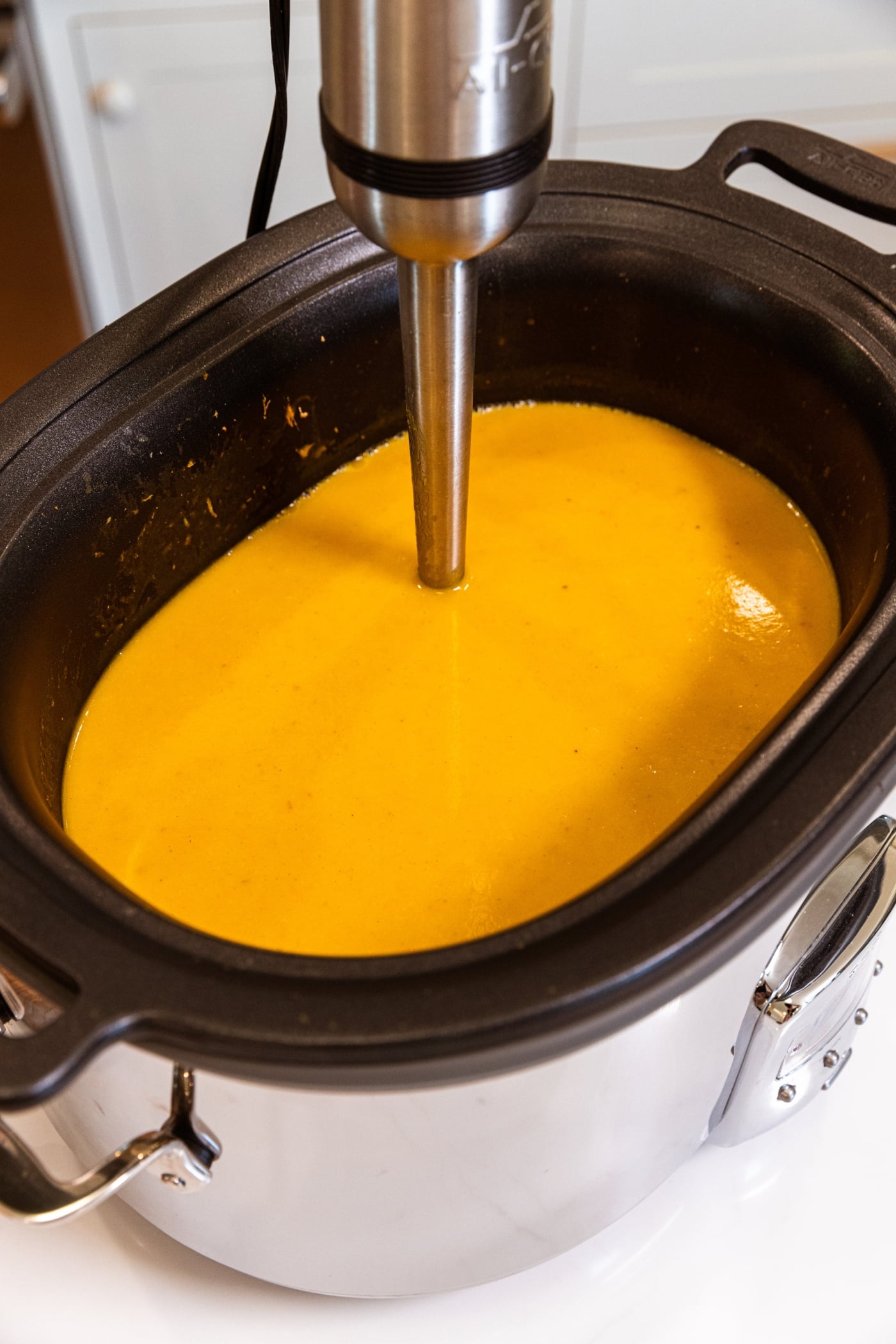 Slow cooker filled with orange colored squash soup with immersion blender finishing blending.