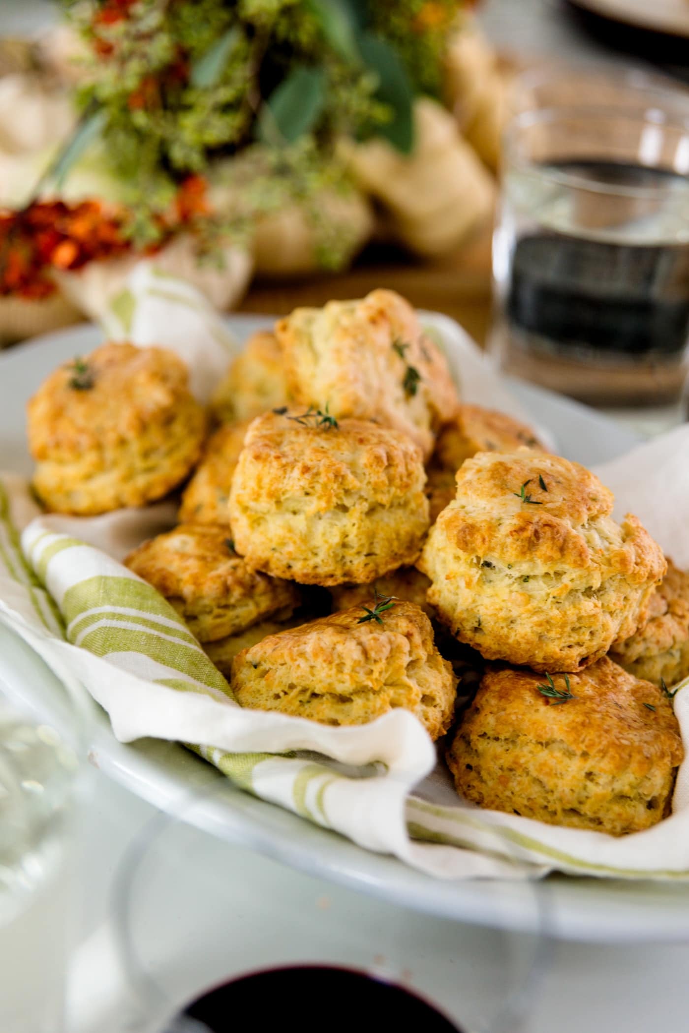 Herb cheese biscuits sitting on white and green linen in white bowl with glasses and flower arrangement in background all on a white surface