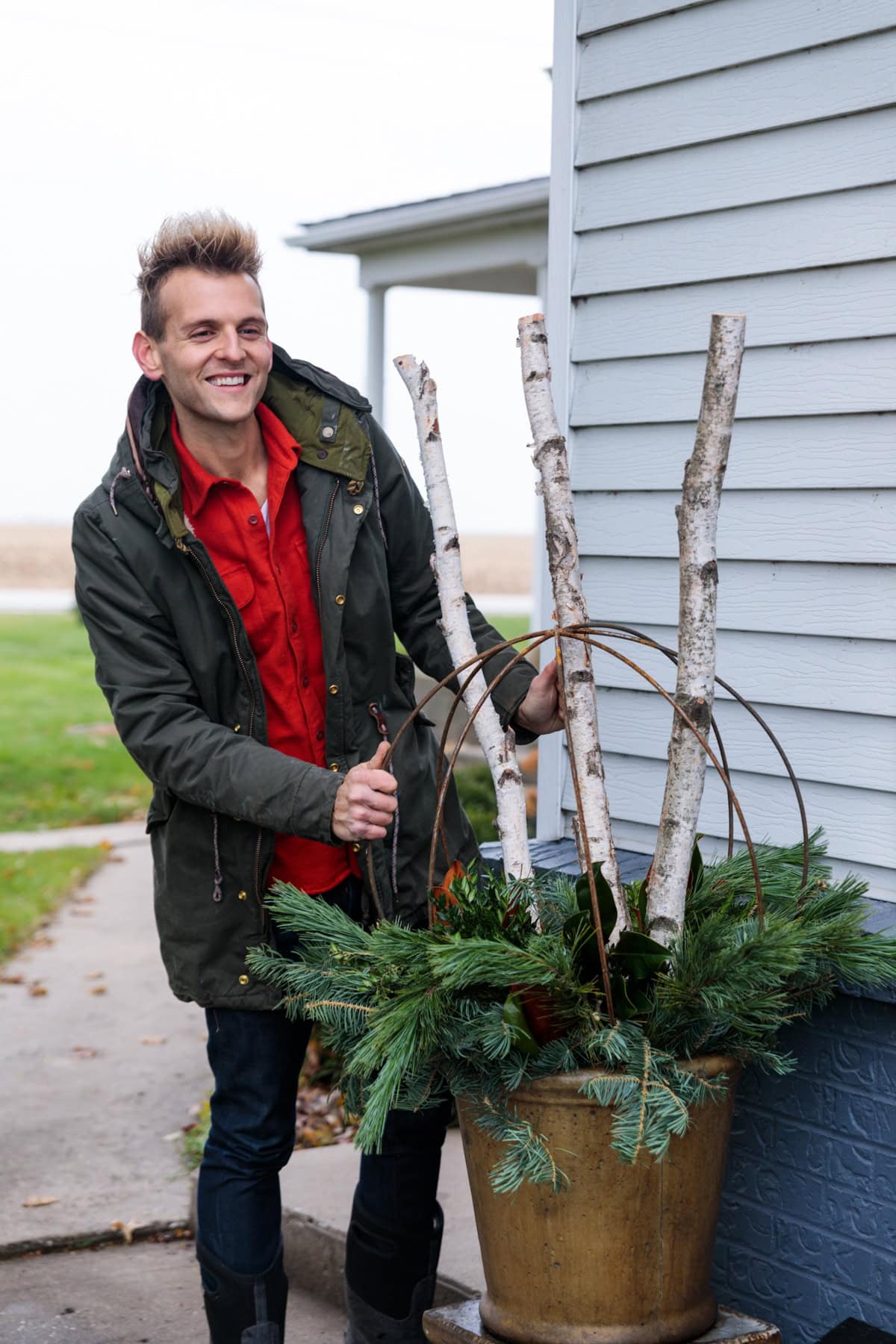 Kaleb Wyse wearing coat and red shirt holding birch sticks in a large urn with a round metal sphere with greens stuffed in amongst as well.