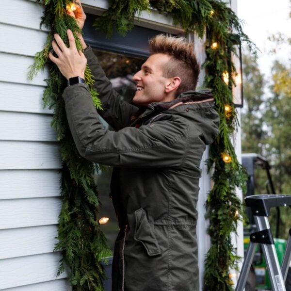 Kaleb Wyse wearing coat hanging Christmas garland with magnolia leaves and lights intertwined around doorway.