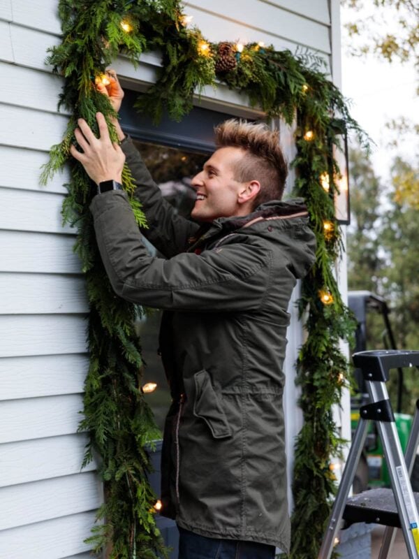 Kaleb Wyse wearing coat hanging Christmas garland with magnolia leaves and lights intertwined around doorway.
