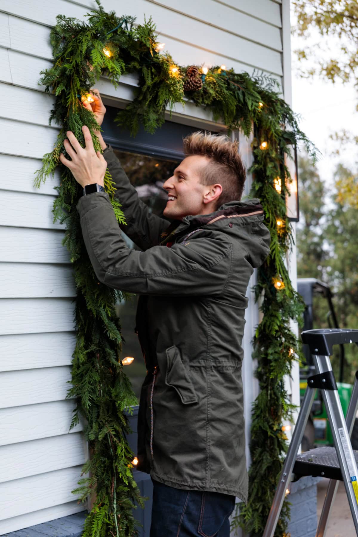 Kaleb Wyse wearing coat hanging Christmas garland with magnolia leaves and lights intertwined around doorway.