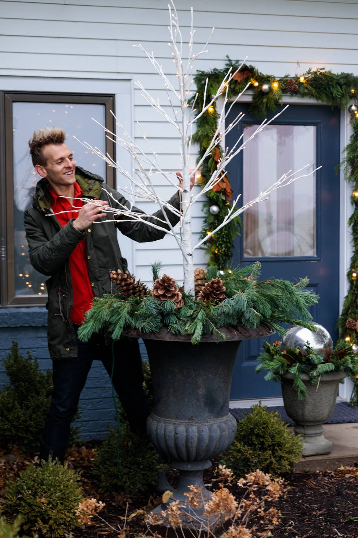 Kaleb Wyse wearing red shirt and coat placing a white tree with small lights on it into a large black urn with greens and pinecones all around the base.