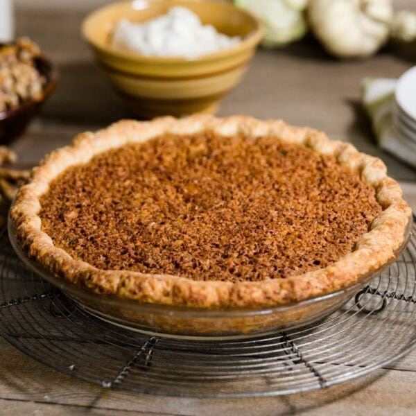 Oatmeal nut pie sitting in glass pie dish on top of cooling rack with white plates, walnuts, sugar and white pumpkins in background on wood plank surface