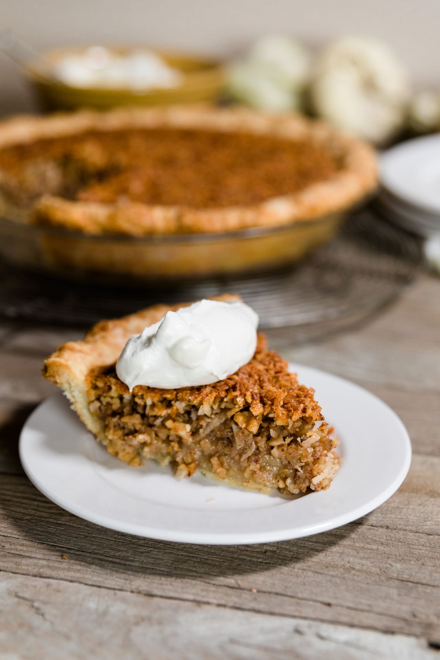 Slice of oatmeal nut pie sitting on white plate with whipped cream on top with rest of pie sitting on wire cooling rack in background along with white pumpkin and extra plates on a wood plank surface