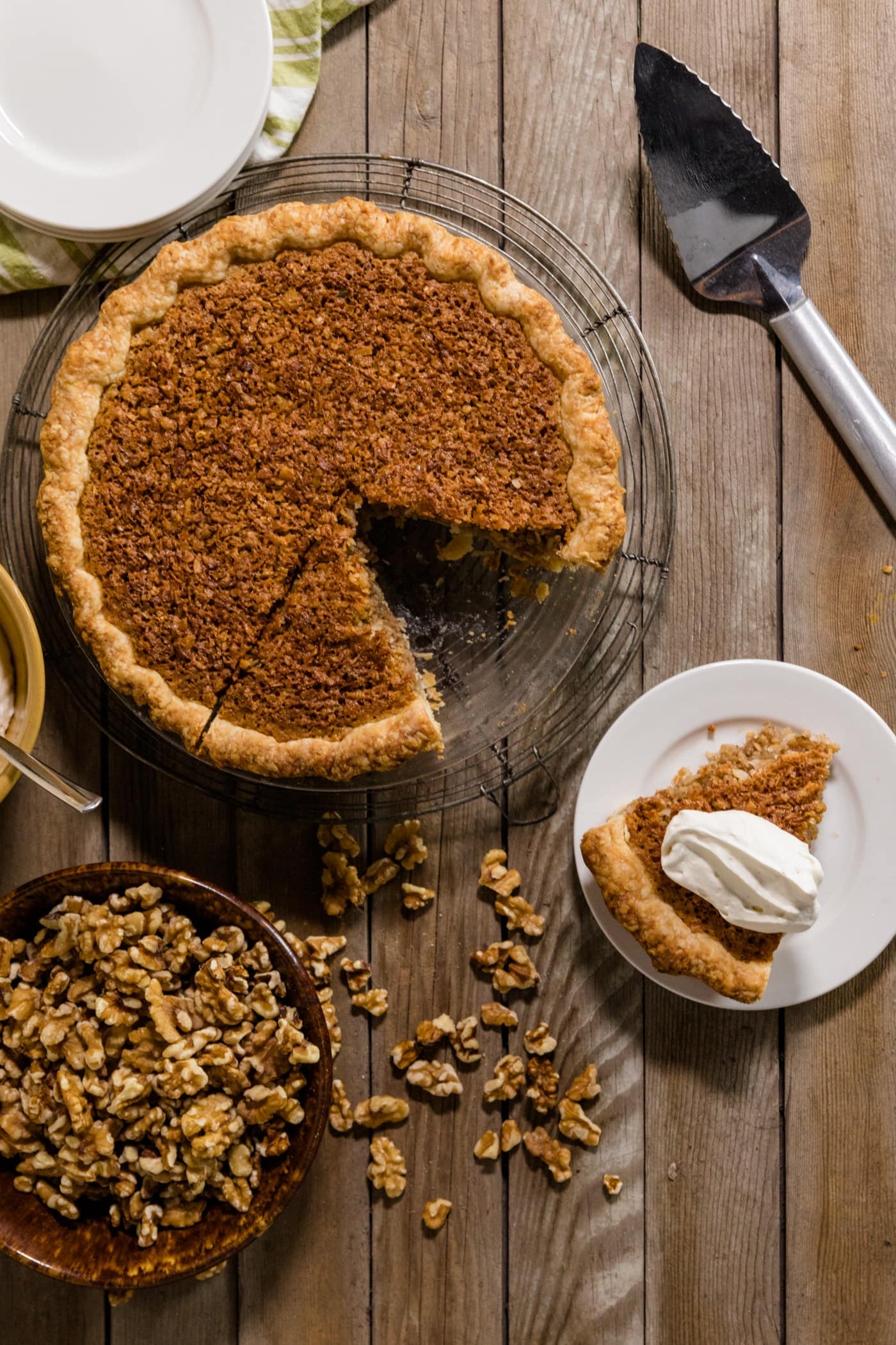 Top down view of slice of oatmeal nut pie sitting on white plate topped with whipped cream with rest of pie, pie slicer, extra plates and toasted walnuts on wood plank surface