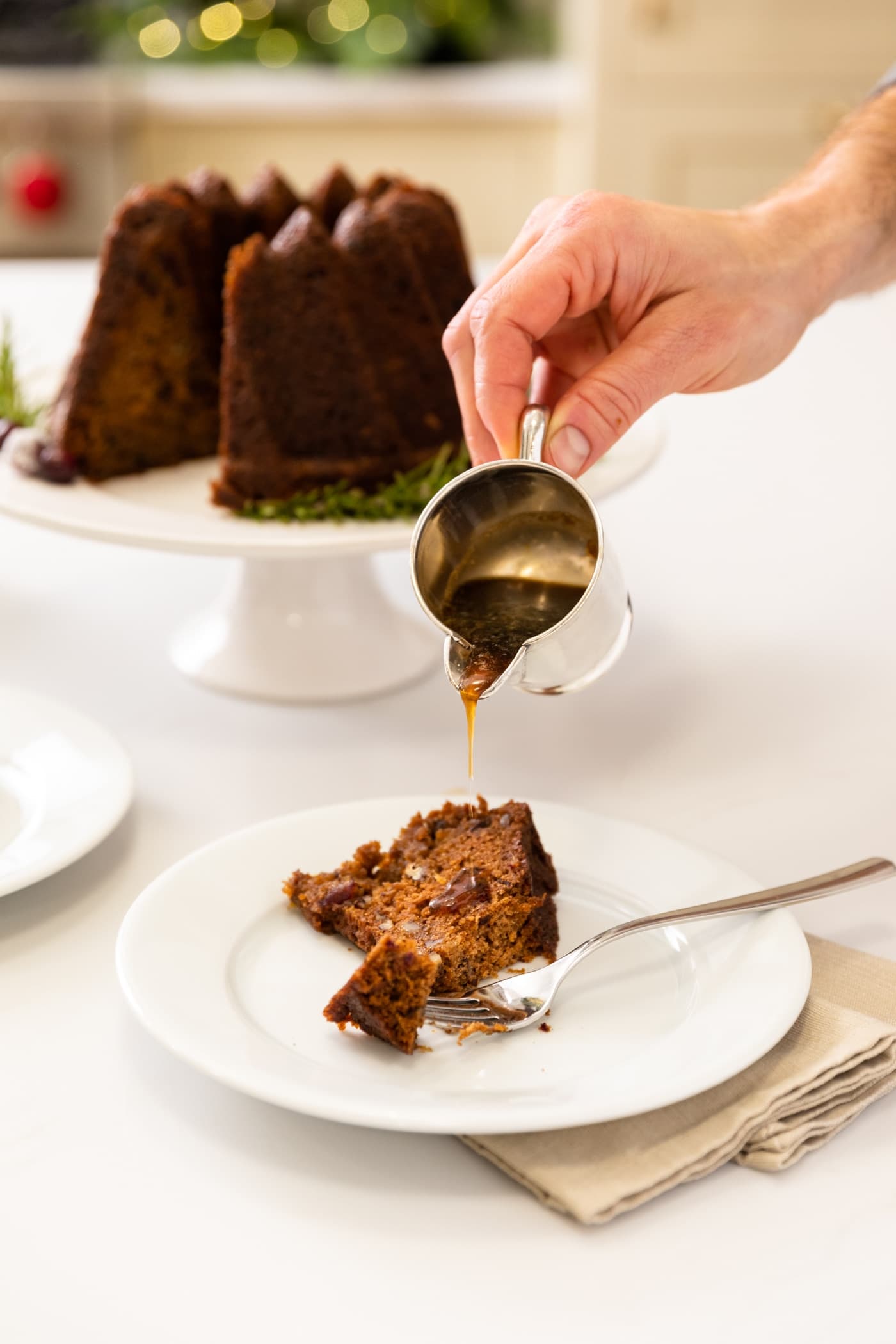 Hand pouring sauce onto piece of brown cake sitting on white plate with rest of cake in background all on white surface