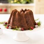 Tall brown cake sitting on white cake plate with cranberries sprinkled around all on white surface with oven and cabinetry in background