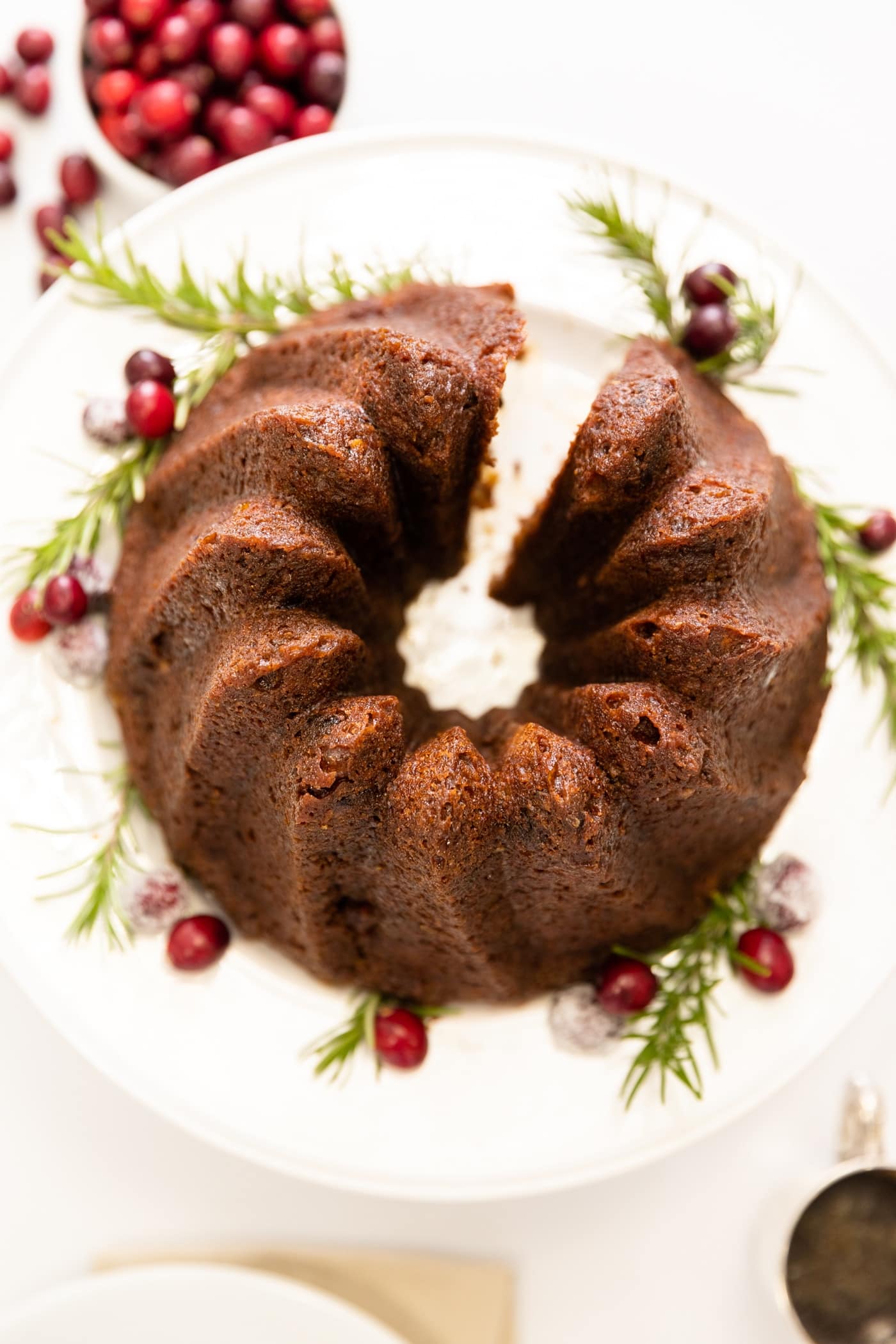 Top down view of brown cake made in Bundt pan sitting on white cake plate with cranberries sitting in bowl and piece of cake to side all on white surface