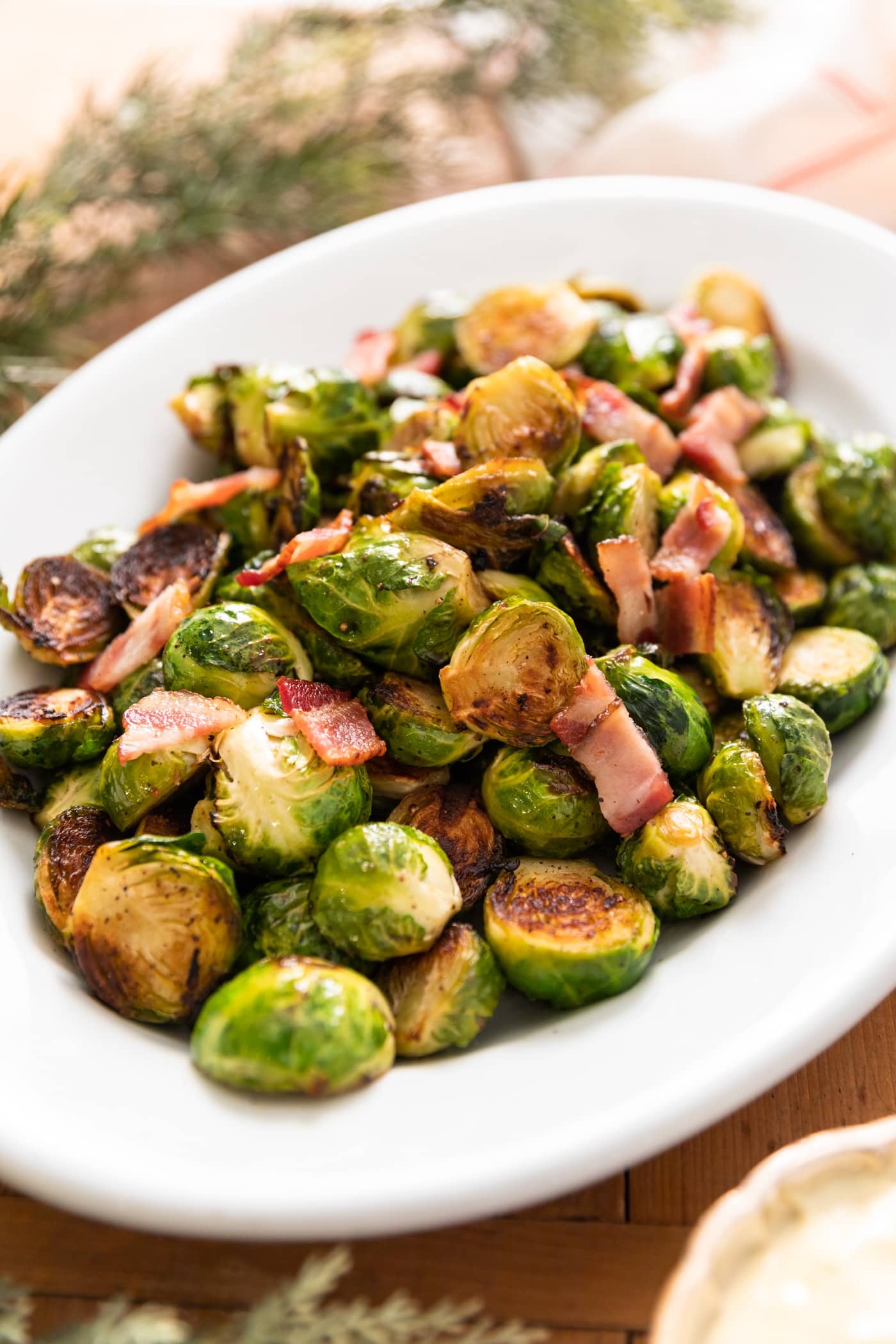 White platter filled with Brussels sprouts and topped with bacon with greenery in background all on wood board