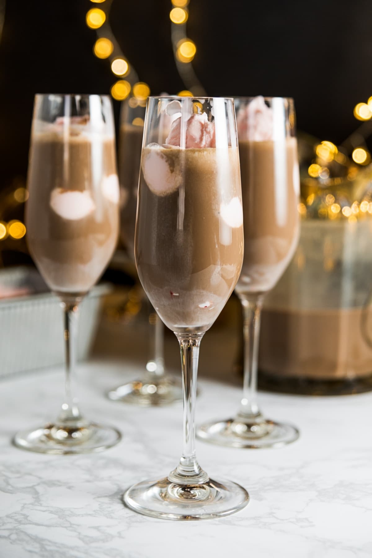 Three champagne flutes filled with pink peppermint ice cream and chocolate coffee mixture sitting on marble surface with Christmas lights on black background