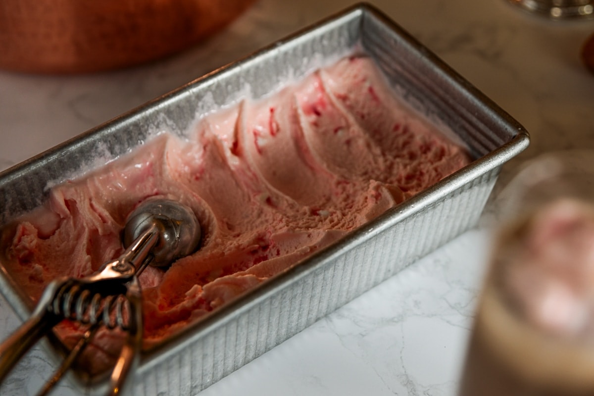 Rectangular metal container filled with pink peppermint ice cream with ice cream scoop resting on top all on marble surface