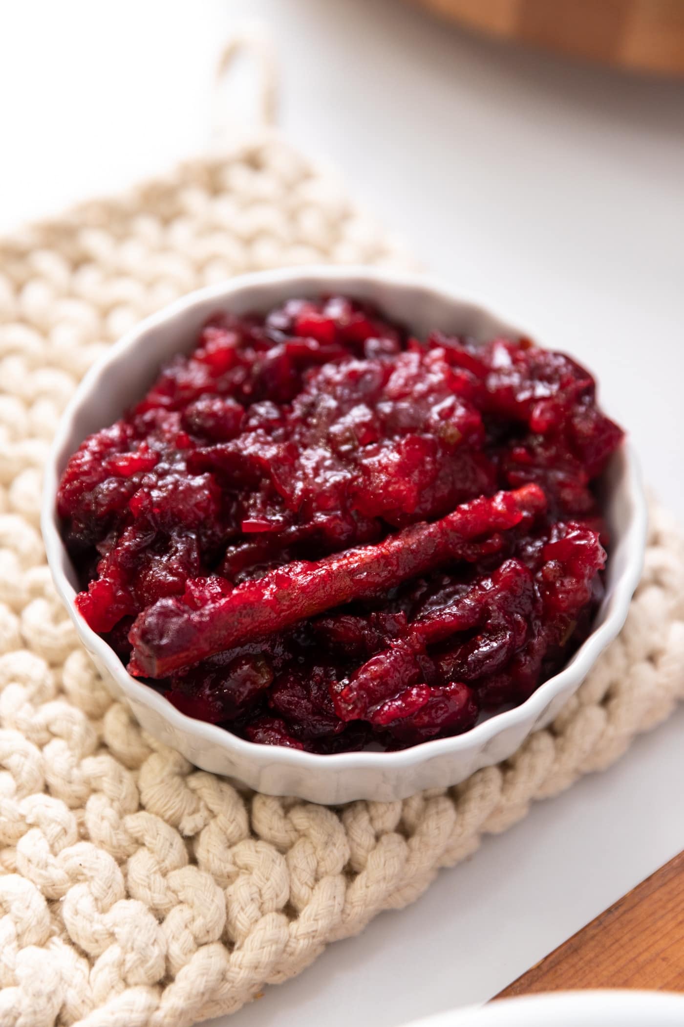 Close up view of red cranberry sauce with cinnamon stick all in white bowl sitting on off-white pot holder on white countertop