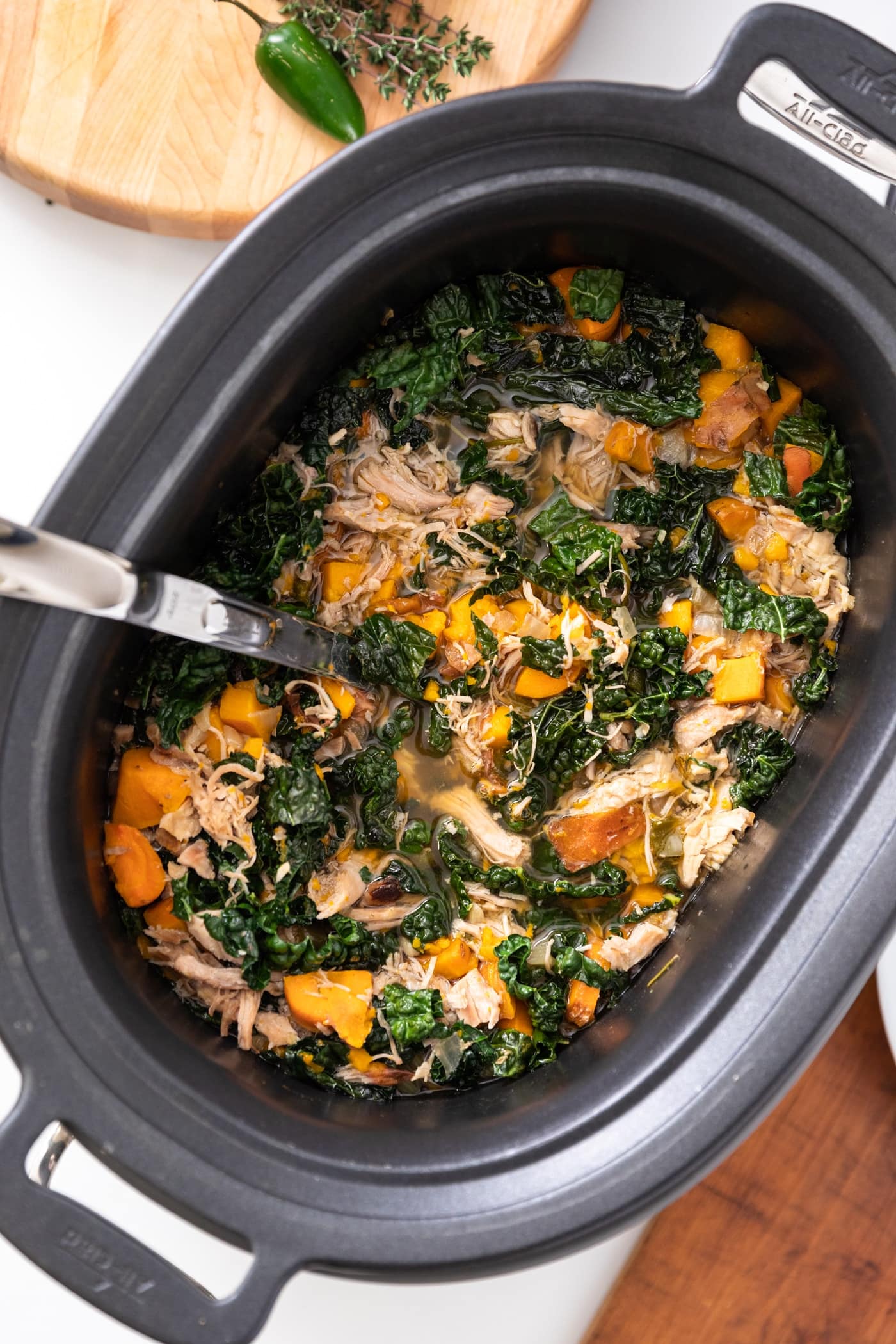 Top down view of turkey soup sitting in slow cooker with kale and sweet potatoes and ladle resting against side all on white countertop surface