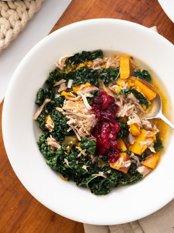 Top down view of slow cooker turkey soup served in a white bowl with soup spoon on the side amidst pieces of kale and sweet potato and topped with a cranberry sauce sitting on wood board with napkin underneath