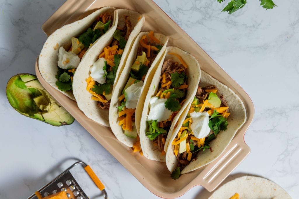 Five tacos in flour tortillas with slow cooker carnitas meat on marble surface.