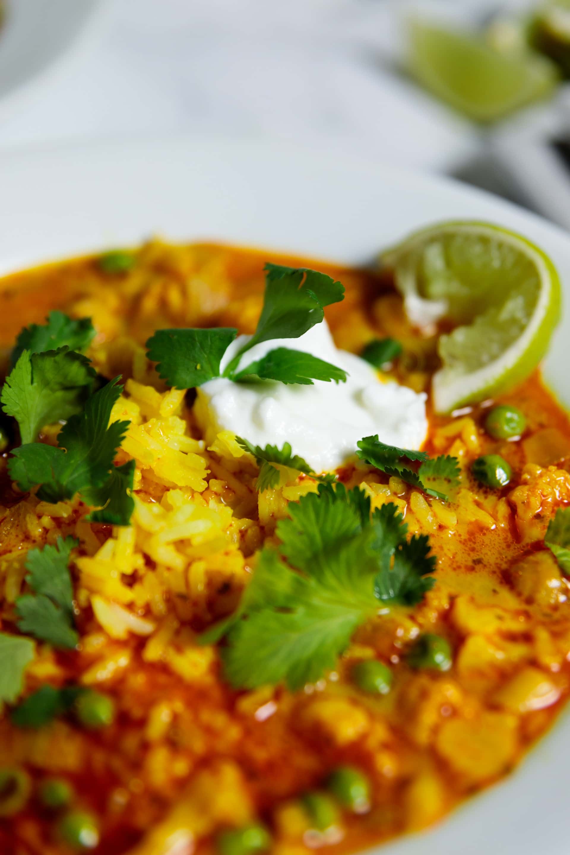 Close up view of orange colored cauliflower curry soup sitting on marble surface topped with cilantro pieces and slice of lime with dollop of sour cream
