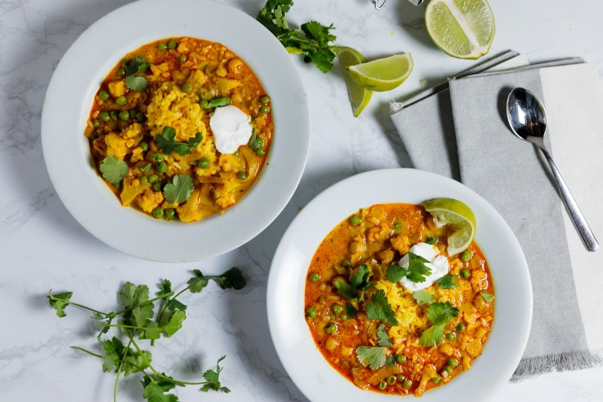 Two bowls filled with orange colored slow cooker cauliflower curry soup sitting on marble surface topped with cilantro and lime slices