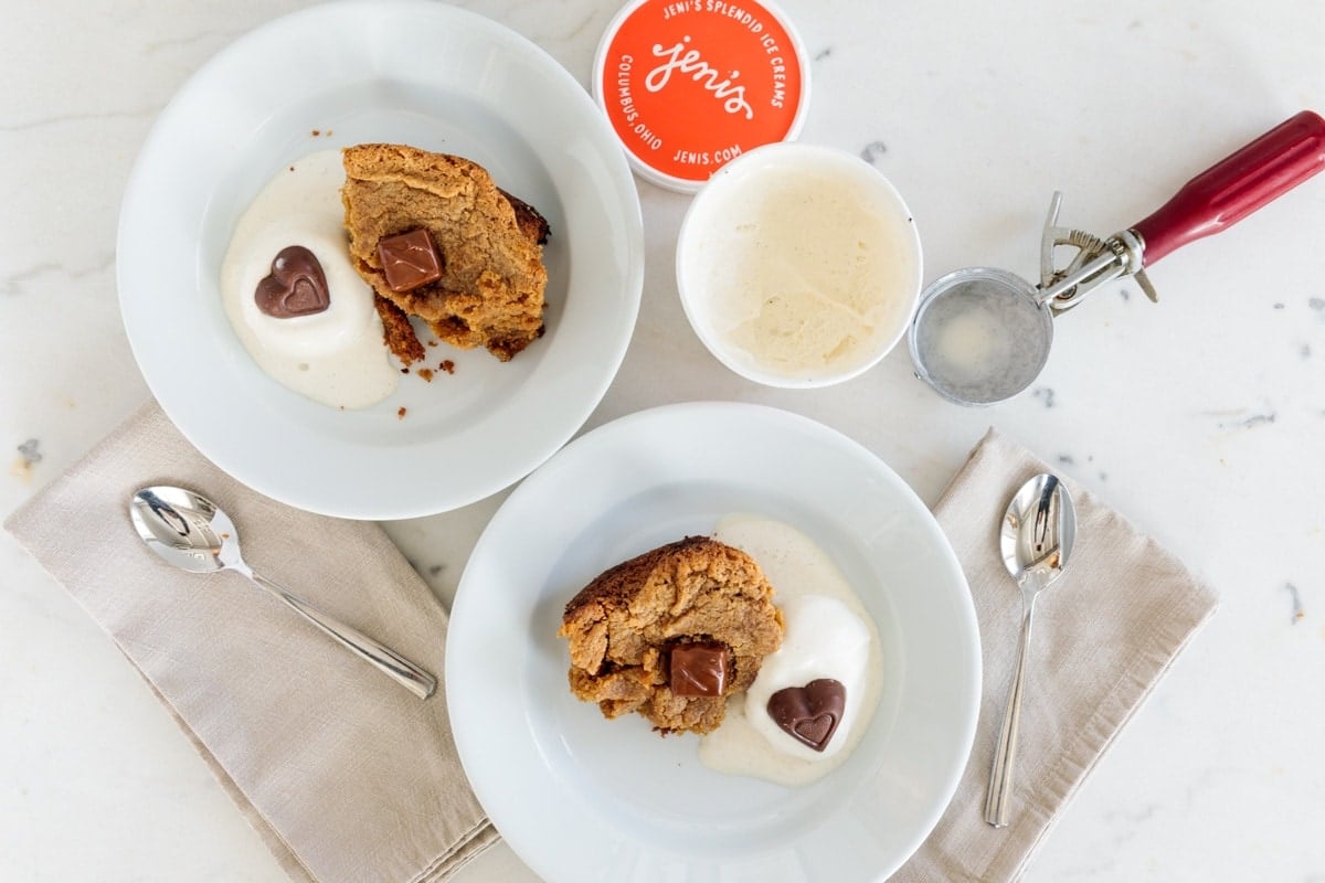 Two plates with pieces of cake with scoop of ice cream on each plate melting slightly all on white marble surface.