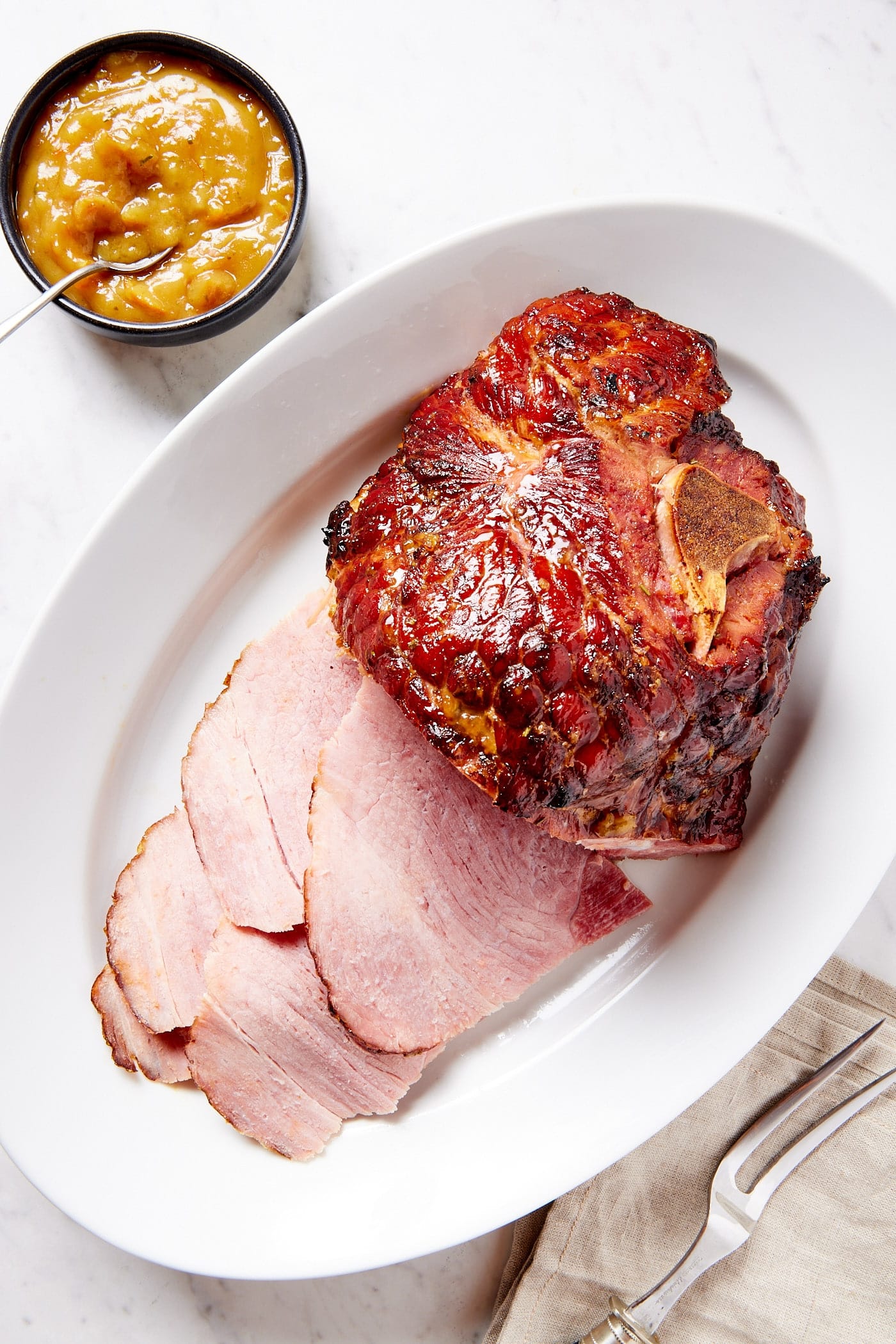 White oval serving platter with ham with slices laying to the side along with orange glaze in small bowl.