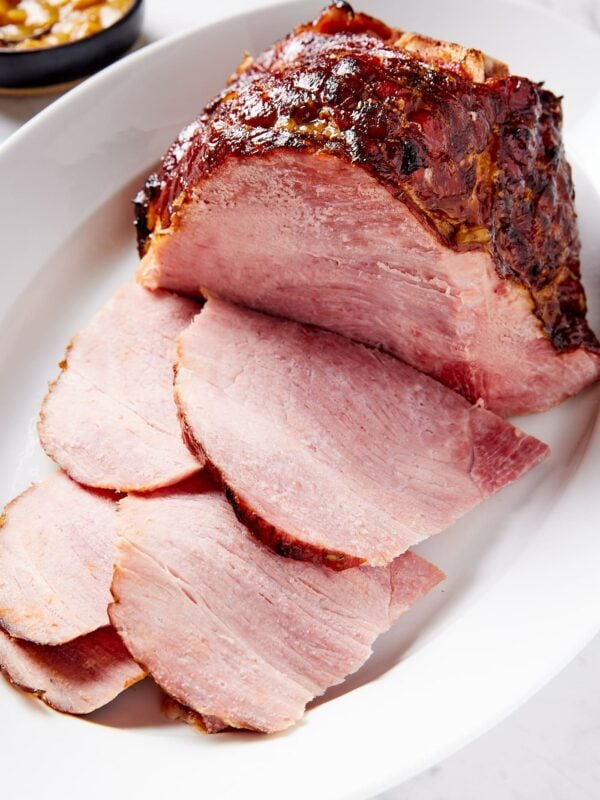 White serving platter with slices of ham piled with rest of ham behind and a small bowl of orange glaze as well.