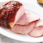 White oval platter filled with slices of ham with rest of ham in the background along with small black bowl filled with orange marmalade glaze.