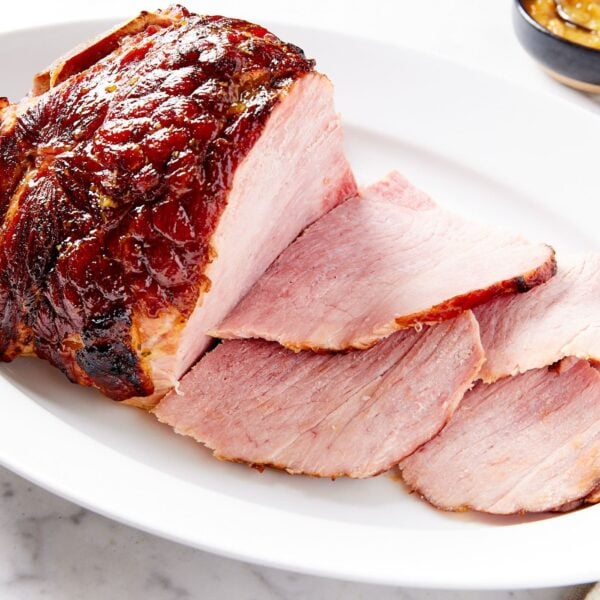 White oval platter filled with slices of ham with rest of ham in the background along with small black bowl filled with orange marmalade glaze.