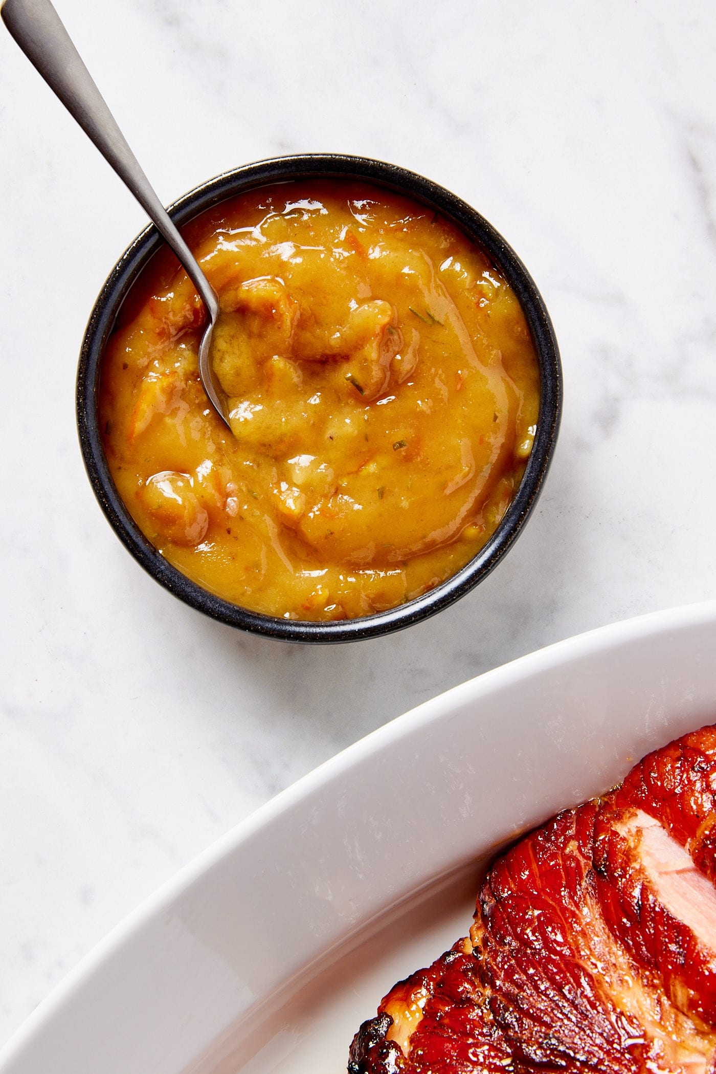 Small black bowl filled with citrus glaze with spoon resting inside with ham to the side.