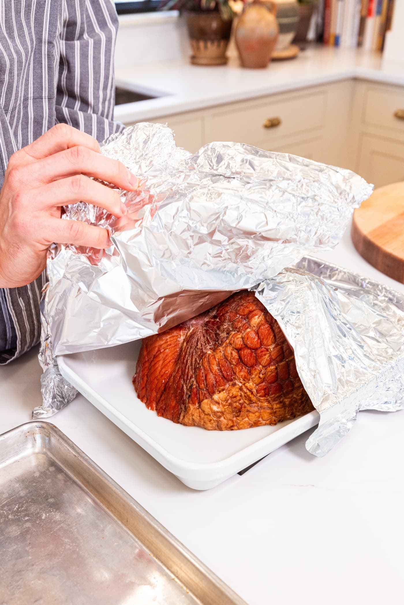 Two hands placing tin foil over the top of a ham with the cut side down in large white baking dish.