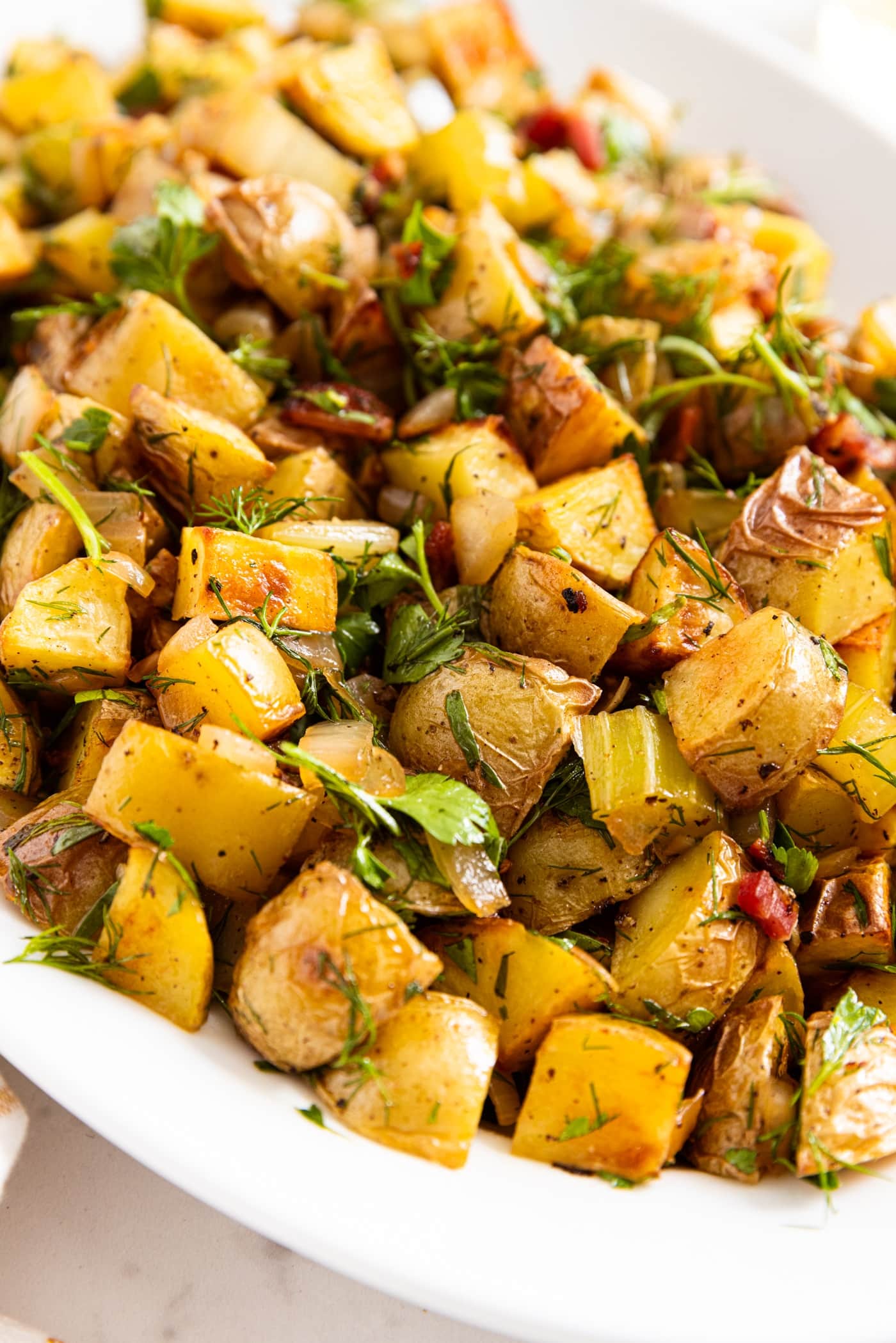 White serving platter with roasted German potato salad sitting on white marble surface with chopped herbs all over the top along with bacon.