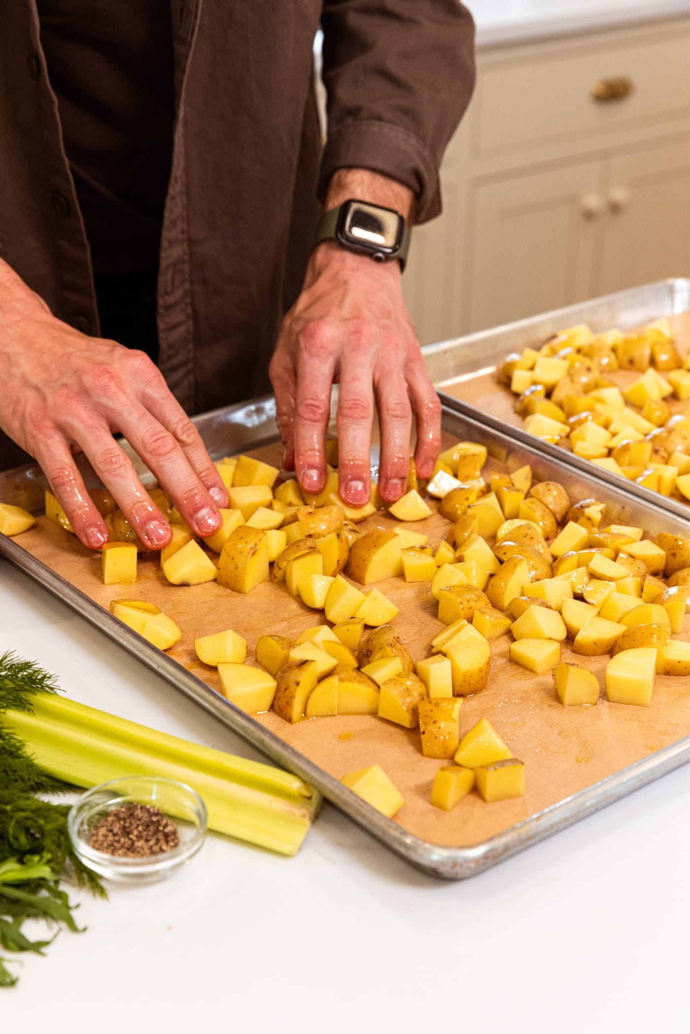 Two hands spreading diced potatoes into an even layer on parchment-lined baking sheet with olive oil drizzled on top all on white surface.