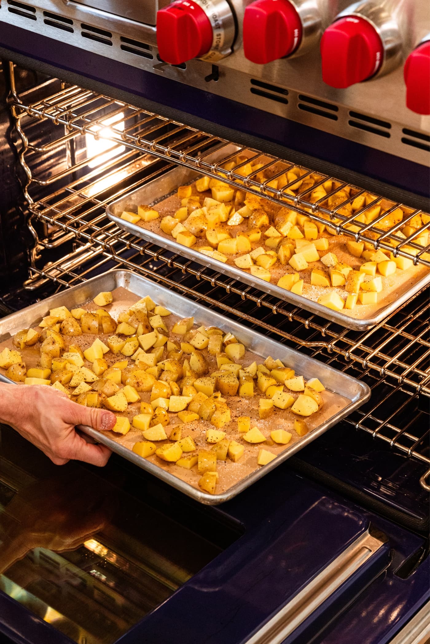 Hand placing one baking sheet with diced potatoes into an oven with a baking sheet already in the oven roasting.