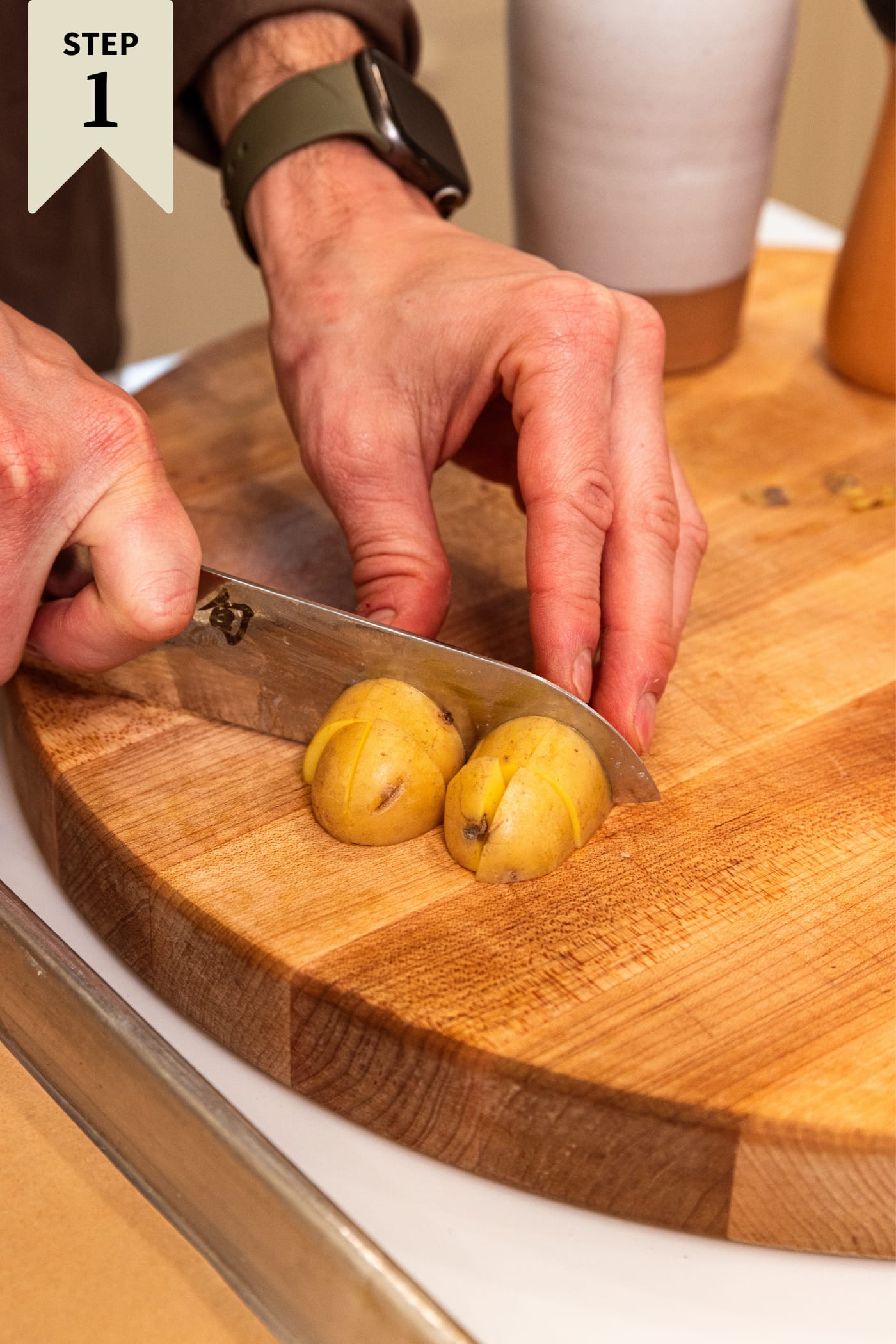 Hand using knife to slice small yellow potatoes with skin still on in half on wood cutting board.