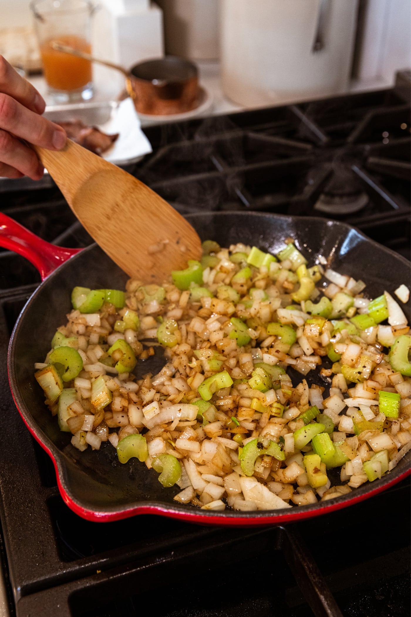Hand holding wooden spatula stirring together browned diced onion and celery mixture in red skillet on stovetop.