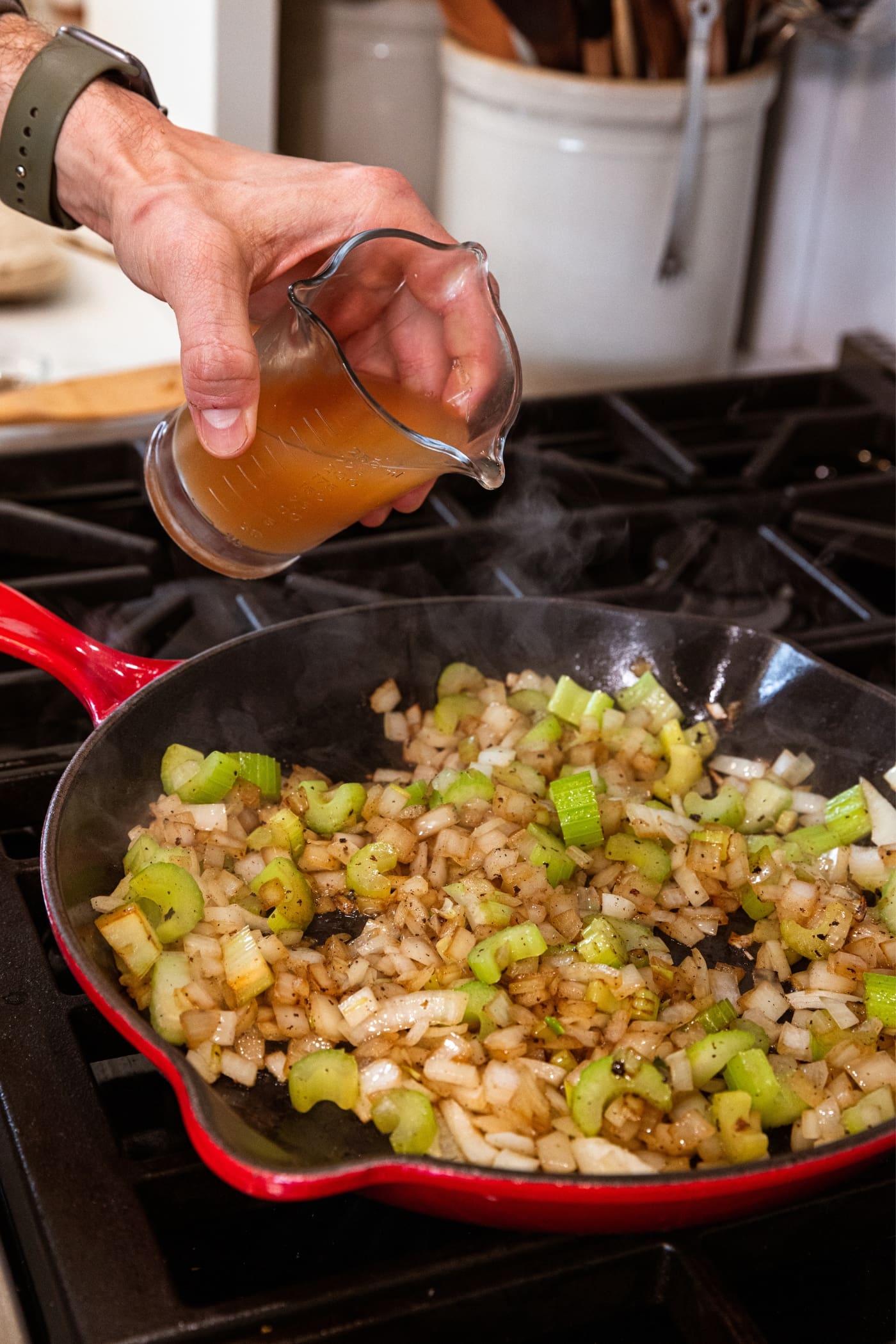 Hand holding glass measuring cup filled with apple cider vinegar about to pour into a red skillet with browned diced onion and celery.