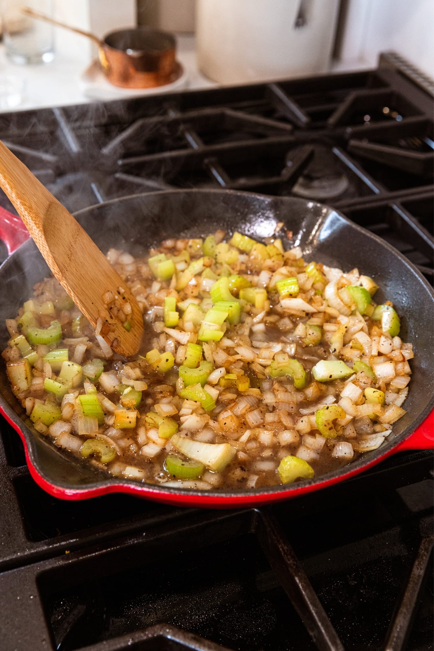 Wooden spatula stirring together simmering apple cider vinegar mixed with chopped onion and celery in red skillet.
