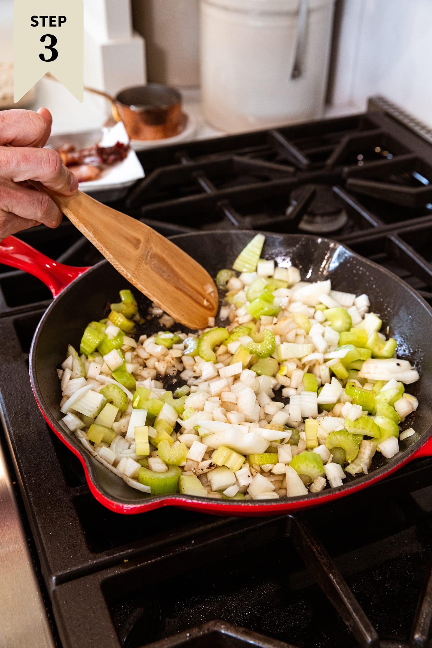 Hand holding wooden spatula mixing together onion and celery together in red skillet.