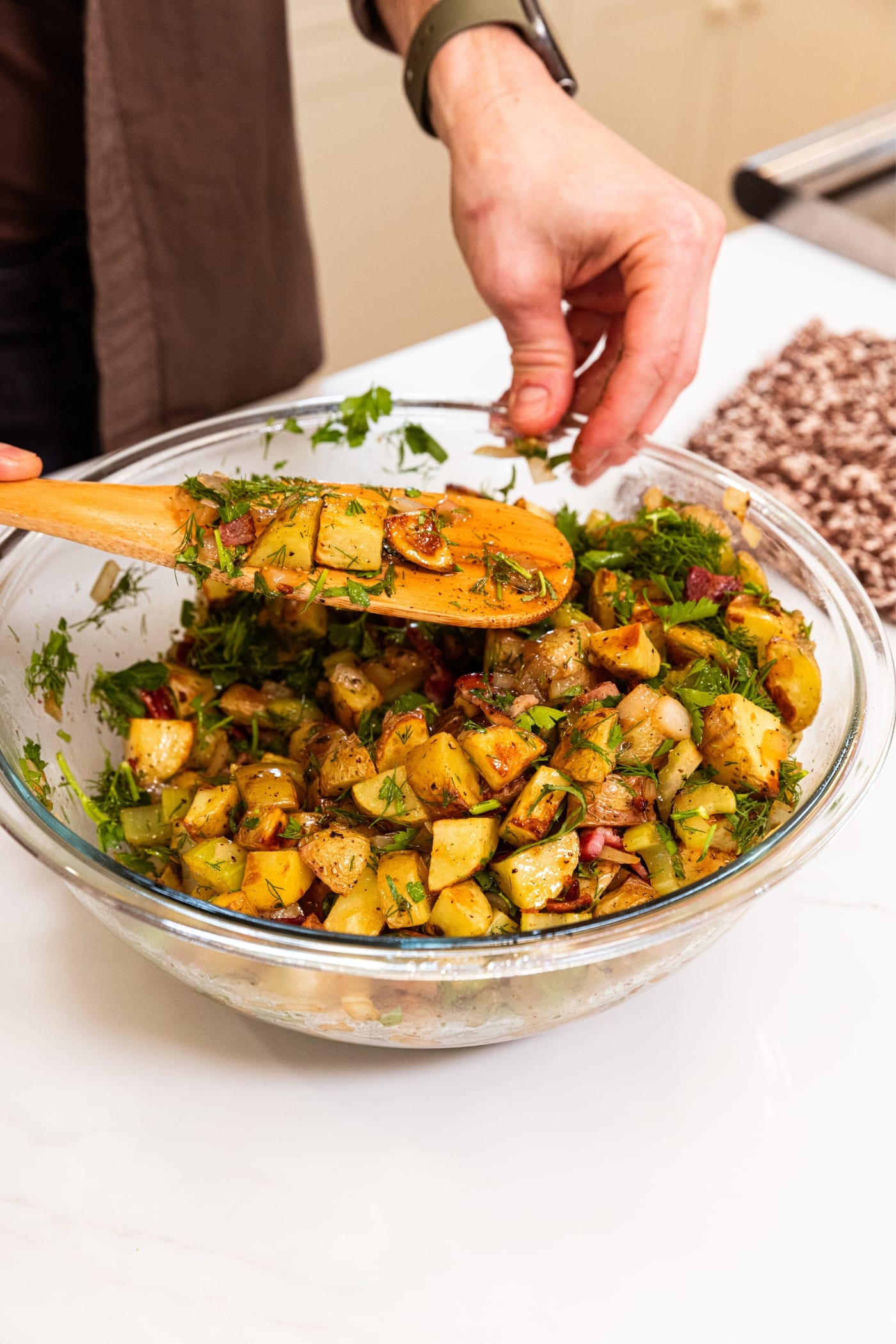Wooden spoon mixing together roasted potatoes with herbs and diced onion and celery mixture on white surface.