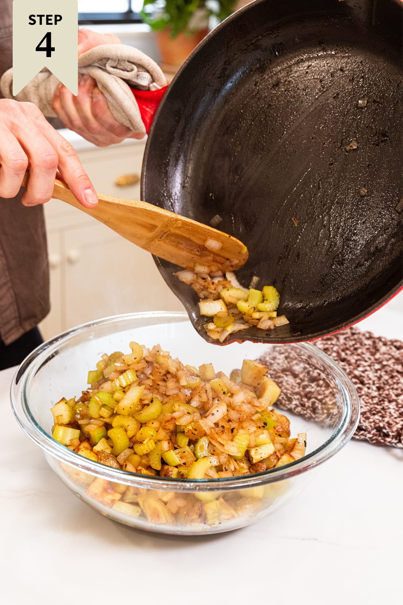 Hand holding red skillet pouring diced onion and celery on top of roasted potatoes in large glass bowl on white surface.