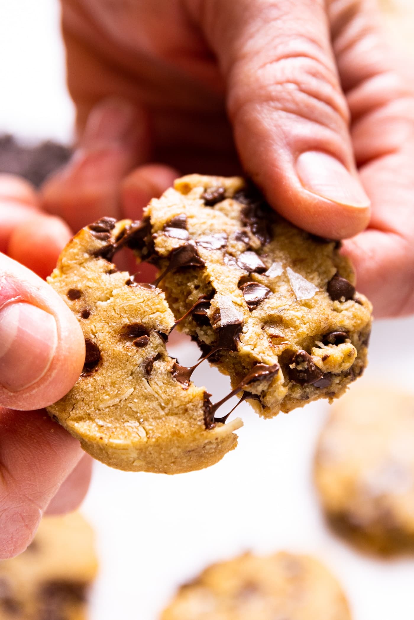 Two hands holding chocolate chip cookie being pulled in half with pieces of chocolate clinging to each side of cookie