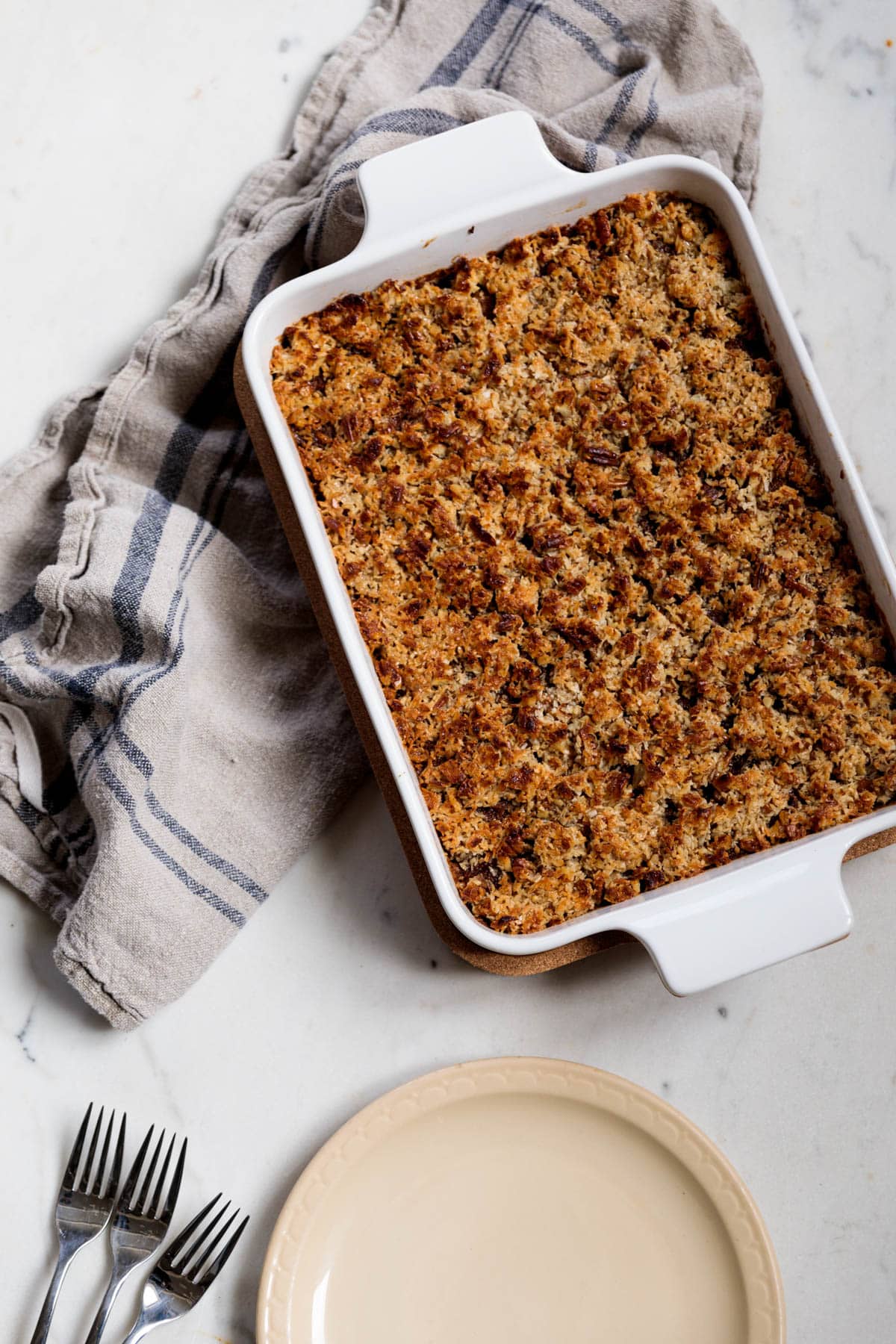 White baking dish with oatmeal cake with tan plate and towel to the side.