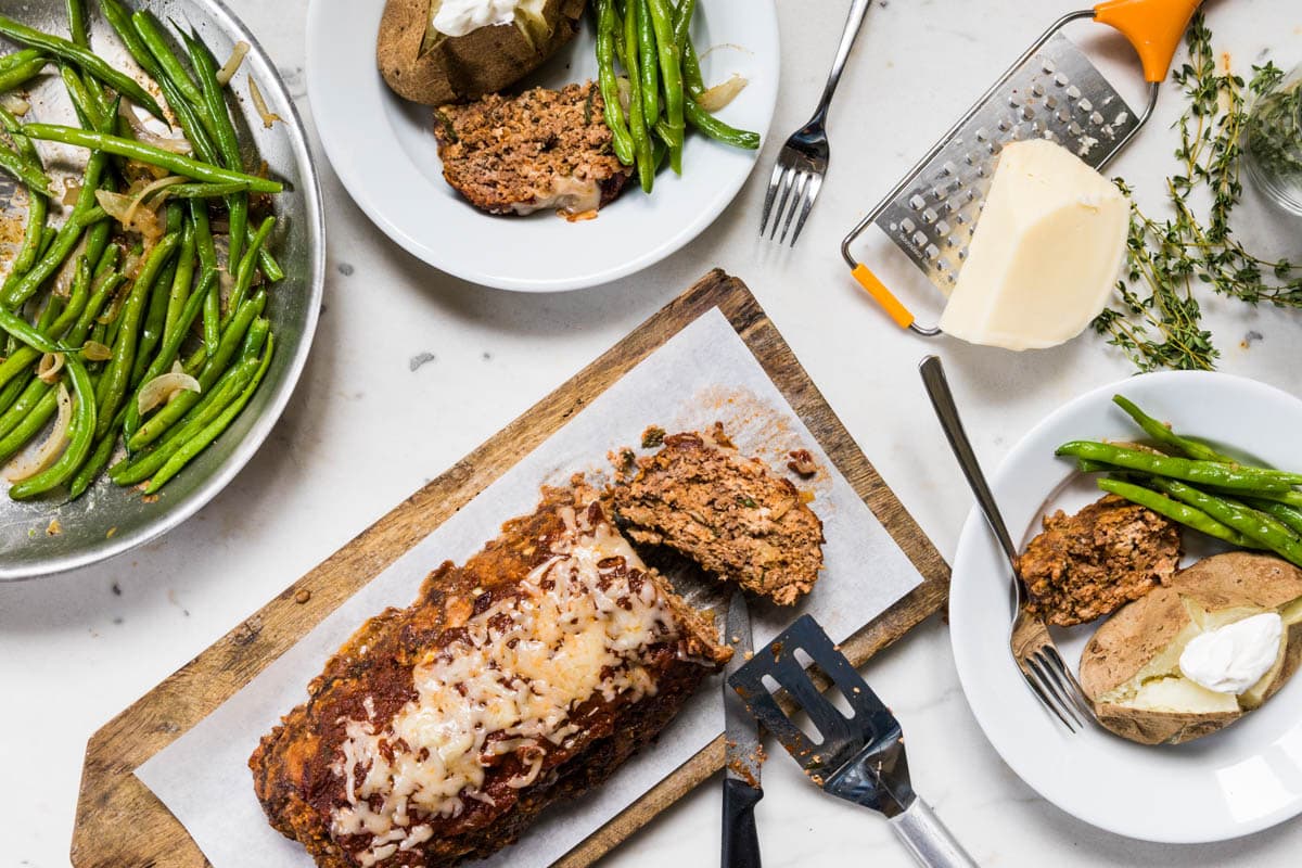 White marble surface with green beans, plates with slices of meatloaf and baked potatoes, and rest of meatloaf on cutting board.