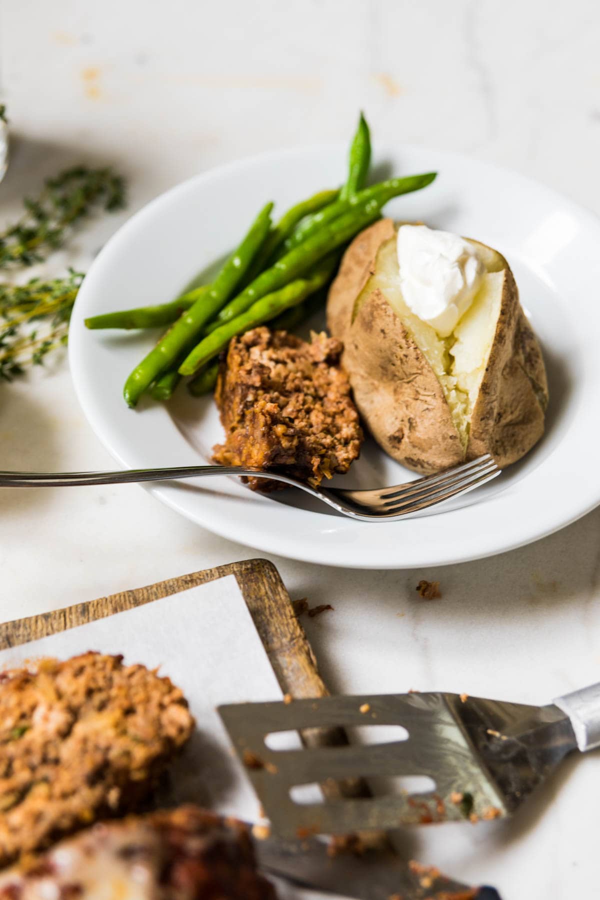 White plate with baked potato, green beans, and a slice of pizza meatloaf with fork as well.