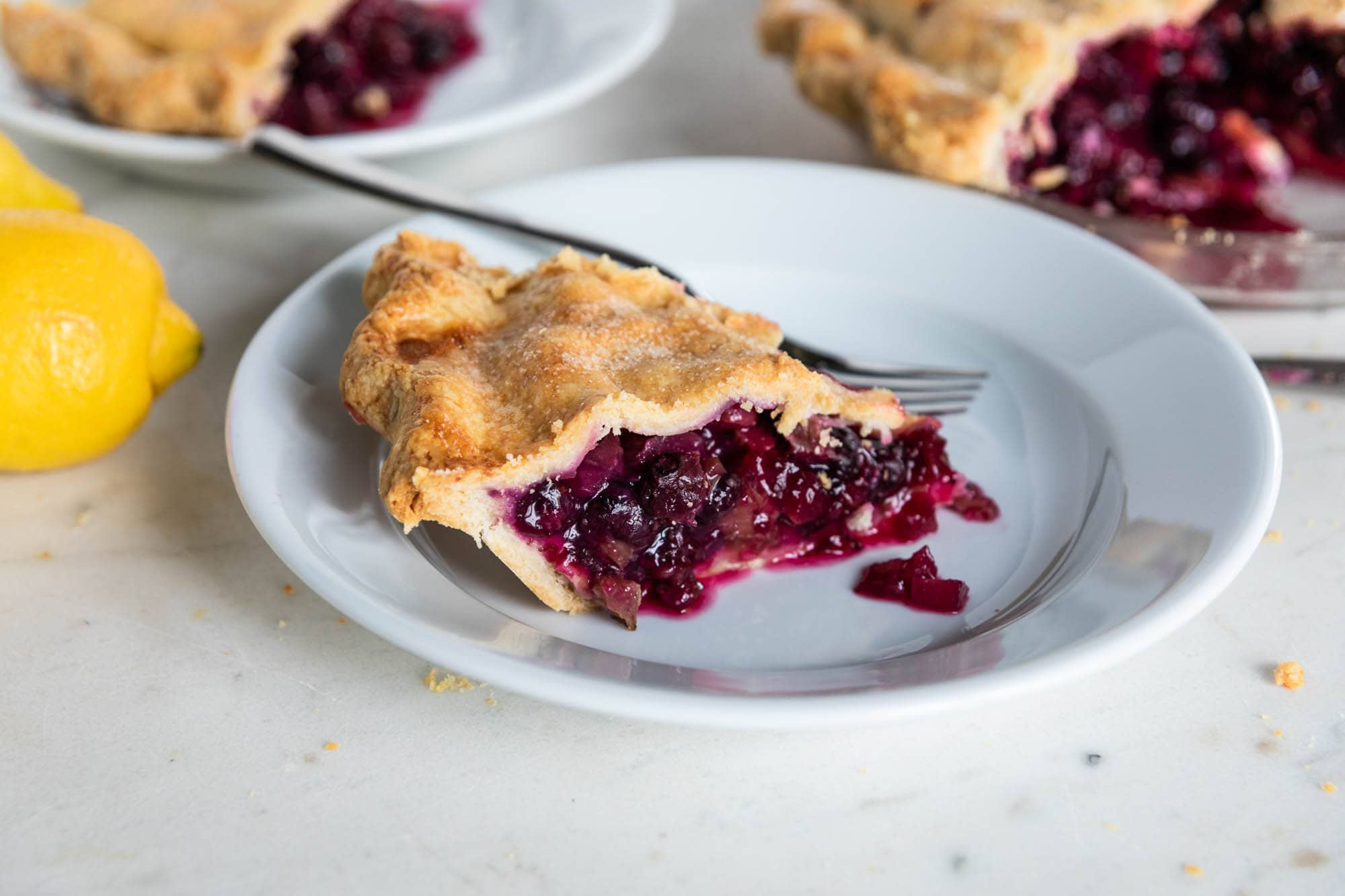 White plate with slice of rhubarb blueberry pie with perfectly baked crust with rest of pie to the side.