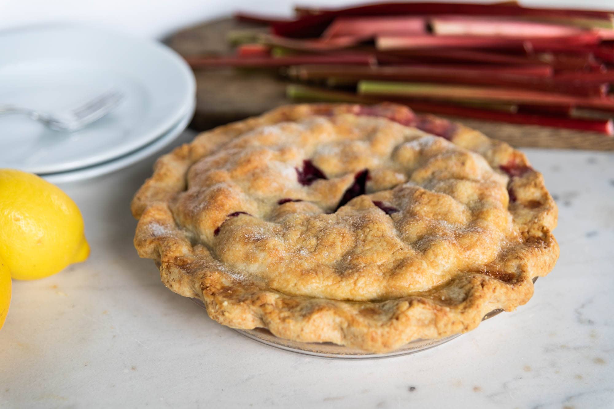 Whole pie after being baked with sugar sprinkled on the top with stalks of rhubarb in the background.